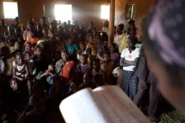 An African woman reading a Bible to a crowded room of listeners