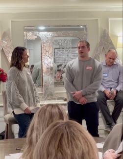 Man standing and speaking with a woman and a man sitting behind him