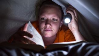 Young woman reading book under covers with flashlight