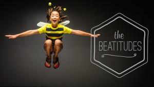 Young child in bee costume in midair, grinning, with the words "The Beatitudes" in the background.