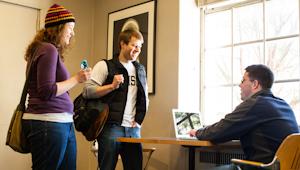 Male and female student walking up to campus minister at table with computer